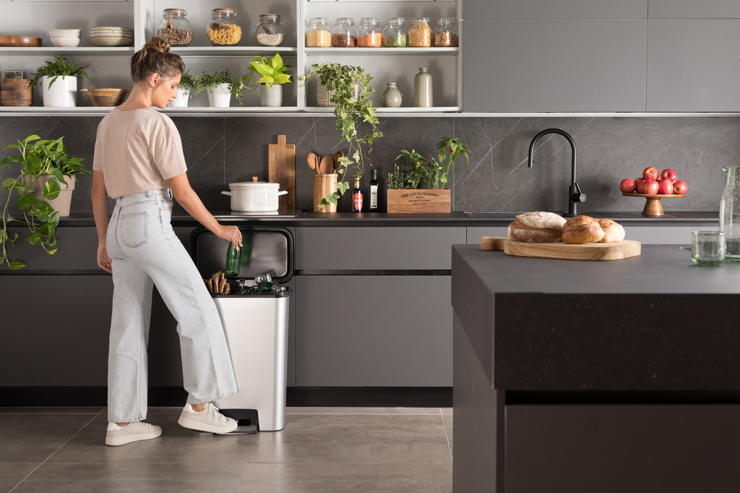 Nő szelektív hulladékgyűjtőbe dob üres üvegpalackot egy modern, növényekkel díszített konyhában.