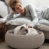 Kép 3/3 - Egy kényelmes, kötött textúrájú kisállat kuckó, világos bézs színben, melyben egy szép macska pihen. Az kuckó mellett egy nő relaxál, a háttérben egy világos szoba és ágy látható.