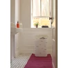 Kép 7/7 - Modern, fehér CURVER 'RIBBON' szennyestartó 49 L, elegáns dizájnnal, lyukas oldalakkal a szellőzéshez, kényelmes fogantyúkkal, fürdőszobai környezetben, rózsaszín szőnyeg mellett.