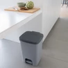 Kép 3/5 - Szürke pedálos CURVER 'ESSENTIALS' szemetes, 20 literes kapacitással, konyhai környezetben található, egy fehér asztal mellett. A design modern és letisztult, a használat kényelmét pedig a nagy pedál biztosítja.